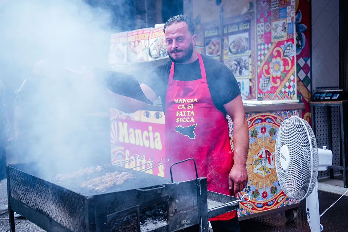 A vendor in a red apron grilling over smoke at a Palermo street food stall